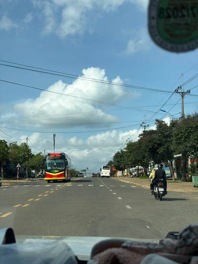 The image depicts a view from inside a vehicle, possibly a car, traveling on a road in Măng Đen, Vietnam. A red and yellow bus with the words "TAN ANH" written on the front is visible on the road, heading in the same direction as the vehicle. A motorbike with two people on it is seen further up the road. Utility poles and wires line the side of the road. The sky is blue with white clouds. Trees and buildings are visible on the side of the road.