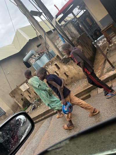 Three young boys are gathered on a paved surface, likely a street or walkway, in Sabojibi, Nigeria. The scene is outdoors, and the ground appears damp, suggesting recent rain or a generally humid environment. Overhead, power lines and corrugated metal roofing are visible, indicating an urban or semi-urban setting. One boy, wearing a dark shirt with red stripes and black pants, is bending down. Another boy, in a navy shirt and tan pants, holds a blue bowl with red markings. A third boy, in a light green garment, is also present. The overall atmosphere seems relaxed, with the boys engaged in their own activities. The lighting is somewhat subdued, consistent with an overcast day. There is no visible text in the image.