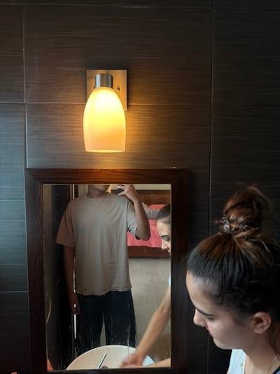 A woman with a bun in her hair peers into a bathroom mirror. Reflected is a full shot of a person with a light-colored shirt and dark pants, seemingly washing their hands in the sink. The scene includes a wall-mounted light fixture with a frosted white shade. The background is a textured gray wall behind the mirror and light.
