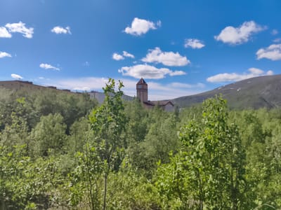 A bright, sunny day with a blue sky dotted with white, fluffy clouds. The scene is a landscape dominated by lush green vegetation, with a dense forest in the middle ground and foreground. In the distance, a large, light-colored building with a distinctive clock tower can be seen peeking through the trees. To the right, a mountainous landscape rises up, with some patches of snow visible on its slopes. The overall impression is one of a natural, somewhat remote setting bathed in sunlight. There is no visible text in the image.
