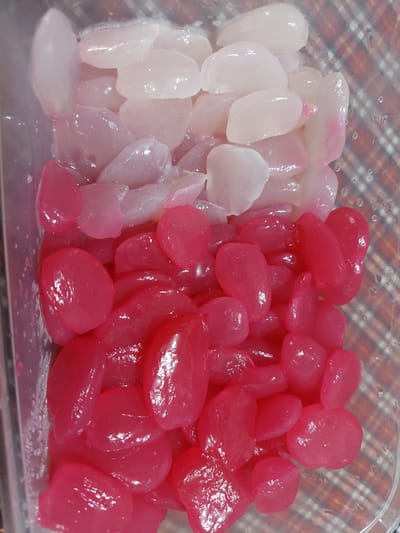 A transparent plastic container holds a serving of kolang-kaling, a type of preserved fruit. The contents are divided into two distinct color sections: the top portion consists of white, translucent kolang-kaling, while the bottom portion features bright pink kolang-kaling. The fruits are oval to teardrop-shaped and appear glossy, suggesting they are preserved in syrup. The container is placed on a surface with a checkered pattern, likely a table or placemat, in a home environment in Jelutung, Indonesia. The background is slightly out of focus, with some water droplets visible on the exterior of the plastic container.