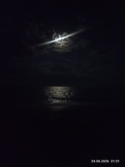 This is a nighttime photograph of a body of water, likely the Caspian Sea, under a cloudy sky illuminated by the moon. The moon is partially obscured by dark clouds, but its light creates a bright, shimmering reflection on the water's surface, forming a path of light that stretches towards the viewer. The sky is predominantly dark, with scattered clouds catching the moonlight.
The scene appears to be a serene and somewhat mysterious landscape. The darkness of the night and the play of light on the water evoke a sense of calm and tranquility. There are no people or man-made objects visible in the frame, focusing the viewer's attention entirely on the natural elements of the sky and water.
The bottom right corner of the image contains a timestamp: "24.06.2026 21:31". This indicates the photograph was taken on June 24, 2026, at 9:31 PM. The location context provided is Шұбартүбек, Kazakhstan, which is known for its proximity to the Caspian Sea. Therefore, the body of water is likely the Caspian Sea. The weather appears to be partly cloudy, typical for a summer night.