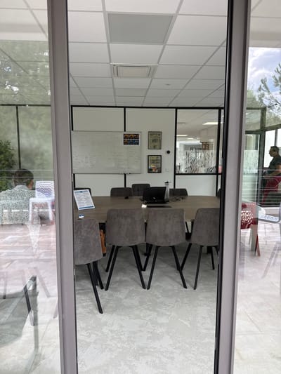 This image, shot from outside a glass window or door, captures an interior meeting room setting in Les Arcs, France. The room features a large meeting table surrounded by upholstered chairs with black metal legs. The backdrop includes a whiteboard filled with mathematical equations and notes, indicating an academic or technical environment. The room's design incorporates tiled ceilings and a glass partition with a frosted pattern. The reflection on the glass shows a person seated, possibly a patron of a café. Beyond the window, glimpses of trees and a clear sky further establish the location.