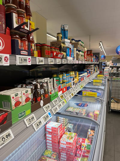 The image shows a grocery store aisle in Köln, Germany (50.919, 6.923). The aisle is lined with shelves stocked with various food items, including sauces, spreads, and frozen foods. On the left side, there are shelves displaying various types of sauces and spreads, primarily packaged in glass jars. The shelves are labeled with price tags. On the right side, a refrigerated unit is stocked with frozen food items packaged in cardboard boxes, also labeled with price tags. The frozen food unit is lit with bright, fluorescent lights. The aisle is well-lit and the shelves are clean and organized. No people are visible in the image.  The environment is sterile and brightly lit, typical of a modern supermarket.