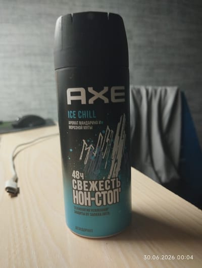 An AXE Ice Chill deodorant spray can is displayed upright on a light-colored wooden surface, possibly a desk. The can is predominantly black with a teal and white label. The label features the AXE logo in large white lettering, followed by "ICE CHILL" in blue. Below this, in smaller white text, are the Russian words "АРОМАТ МАНДАРИНА И МОРОЗНОЙ МЯТЫ" (Mandarin and Mint Aroma). A graphic of stylized ice shards or mountain peaks is present on the lower half of the label. Further down, "48Ч СВЕЖЕСТЬ НОН-СТОП" (48H NON-STOP FRESHNESS) is printed in large white lettering. Beneath this, smaller Russian text reads "ТЕХНОЛОГИЯ УСИЛЕННОЙ ЗАЩИТЫ ОТ ЗАПАХА" (Enhanced Odor Protection Technology). At the bottom of the label, "ДЕЗОДОРАНТ" (Deodorant) is visible. To the left of the can, a white cable with a grey connector is lying on the surface. In the background, a dark, textured wall or surface is visible, and to the right, a blurred grey fabric is draped, possibly covering a chair or furniture. The lighting appears to be artificial, casting shadows on the wooden surface. A timestamp "30.06.2026 00:04" is overlaid on the lower right of the image. The image does not contain any elements or context that would allow for its localization to Zelenogorsk, Russia.