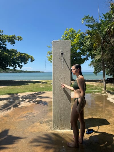 A female subject, viewed in profile from the right, stands barefoot on a wet concrete surface in Rio Bueno, Jamaica. She wears a dark olive green two-piece bikini, and her wet hair is pulled back. Her right hand is extended towards a circular control mechanism on a freestanding, rectangular, light gray concrete outdoor shower column. Water flows from a metal spout on the front of the column, creating a stream that splashes onto the concrete below. Another identical metal spout is visible on the left side of the column. A pair of blue and black sandals rests on the concrete behind the subject. The background features a green grassy area leading to a calm blue body of water, presumably a bay or the sea, with a distant coastline and structures visible under a clear blue sky. Lush green trees and tall palm trees frame the scene, and strong sunlight casts distinct shadows of palm fronds across the concrete ground.