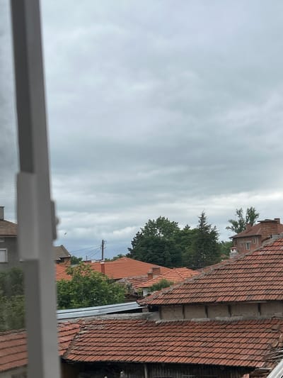 The image shows a view of rooftops in Parvomay, Bulgaria. The sky is overcast with grey clouds. In the foreground, a white window frame is visible on the left. The rooftops are covered with red clay tiles, and some of the buildings have chimneys. Trees are scattered throughout the scene, with a dense cluster of green trees in the middle ground. A few utility poles with wires are also present. The overall impression is of a residential area under a cloudy sky.