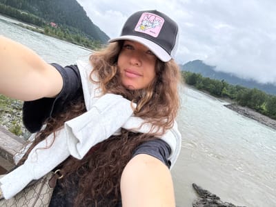 A woman is taking a selfie outdoors near a river with mountains in the background. She is wearing a black and gray trucker hat with a pink patch depicting Daisy Duck, a white sweatshirt draped around her shoulders, and dark gray t-shirt. Her long, curly brown hair is visible. She is holding her arm out to take the photo. The river is wide and appears to be flowing rapidly. The sky is overcast and the mountains are covered in green trees and some clouds. The location appears to be Elekmonar, Russia, given the presence of a river and mountainous terrain.