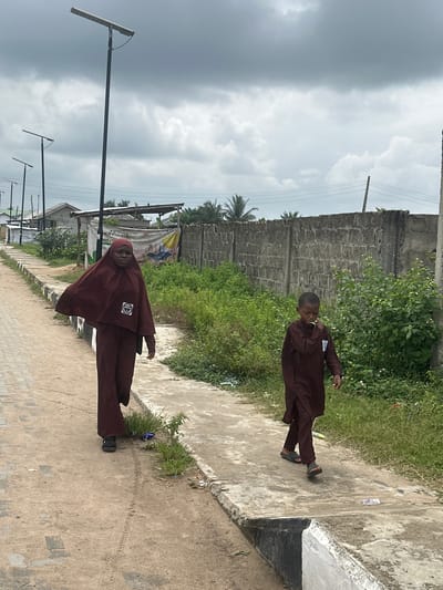 This image, taken in Mosafejaw, Nigeria, depicts an outdoor scene under an overcast sky. Two individuals are walking along a street or path.
On the left, a woman or girl in a maroon full-body covering and hijab walks on a dirt path. A white rectangular patch with a black symbol is affixed to her chest area. Her left arm is slightly extended outwards.
To her right and slightly ahead, a young boy walks on a concrete pavement. He wears a loose-fitting, long-sleeved maroon garment, possibly a tunic or thobe, and sandals. A small white rectangular patch is visible on the left chest area of his garment. His right hand is raised towards his mouth.
The left side of the frame features a dirt path with a visible tire track, bordered by sparse green vegetation and a low curb that separates it from a paved section. Several tall, black solar-powered streetlights ascend into the cloudy sky, extending into the background along the path. Behind these, simple concrete structures and a faded banner with text (partially legible "MARKET," "200") and graphics are visible.
The right side of the frame features a concrete walkway bordered by dense green bushes and smaller plants, some with small white flowers. Behind this vegetation, a tall, grey concrete block wall extends into the distance. Power lines are visible across the upper middle ground against the sky. Palm trees are discernible above the wall in the background. Scattered litter is visible on both the dirt and paved surfaces.