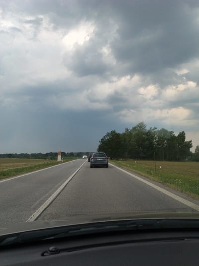 The image depicts a view from inside a vehicle traveling along a road in Plástovice, Czechia. The scene is dominated by a dramatic sky filled with dark, heavy clouds, suggesting impending or ongoing rain. The road is paved and marked with a white line, indicating it's a two-lane highway. To the right of the road, there is a grassy verge with scattered wildflowers and a line of trees forming a dense forest border.
Several vehicles are visible on the road. In the foreground, a dark-colored SUV is driving ahead, its taillights illuminated. Further down the road, another vehicle, possibly a car, is also visible. The perspective suggests the photo was taken from the passenger seat or driver's seat of a moving vehicle, as evidenced by the dark interior frame on the right side of the image.
Along the roadside, there are a few small, white rectangular markers, possibly mile markers or signs, and a distinctive orange and white structure. The overall mood is slightly somber due to the overcast sky, but the presence of the road and other vehicles implies normal travel or commuting. The time of day appears to be daytime, though the cloudy conditions obscure direct sunlight. No people are clearly identifiable within the image. The license plate of the SUV is partially visible, displaying characters that appear to be "E4R 7569".