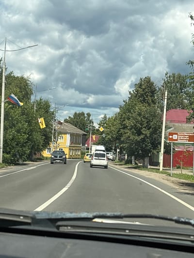 The image shows a view from a vehicle driving on a road in Pereslavl-Zalessky, Russia, under a cloudy sky. On the left side of the road, there are trees and a yellow building. Flags, including the Russian flag, are displayed on poles along the roadside. Several cars are on the road, with one black car in the foreground and a white van and a yellow car further ahead. On the right side of the road, there is a brown sign with text in Russian and English indicating "St. Nicholas Monastery" with an arrow pointing left. There are also trees and a building with a red roof visible on this side. The road has a white dividing line. The perspective suggests the photograph was taken from the driver's or front passenger's seat of a moving vehicle, as evidenced by the dashboard and windshield wipers in the lower part of the frame.