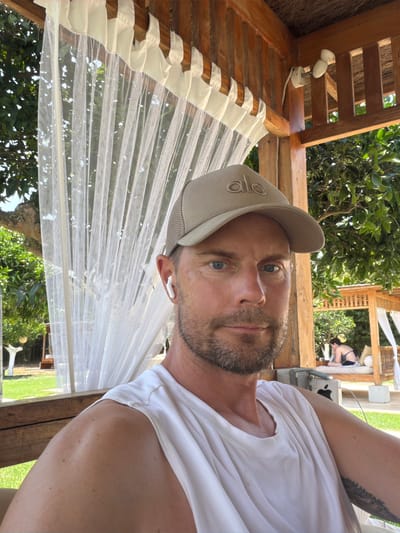 The image shows a man wearing a tan baseball cap and a white sleeveless shirt, posing for a selfie. He has blue eyes and a beard, and he is wearing white earbuds. He is sitting in a wooden structure with a thatched roof, which appears to be an outdoor gazebo or cabana. Behind him, a white sheer curtain hangs, and through it, lush green trees and foliage are visible. In the background, another wooden structure with white cushions and draped fabric can be seen, with a person in a bikini lying on it. The setting suggests a relaxed, possibly resort-like environment, consistent with the city of Sant Joan de Labritja, Spain, known for its natural beauty and tourist accommodations.