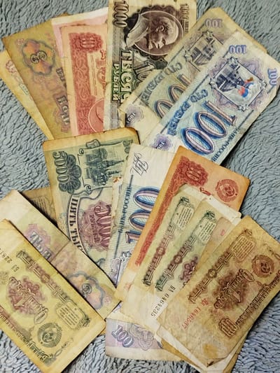 The image shows a collection of old Soviet and Russian banknotes scattered on a soft, textured, gray surface. The banknotes are of various denominations, including 1, 3, 5, 10, 100, 1000, and 5000 rubles. Some of the banknotes feature portraits of Lenin and other Soviet symbols. The overall condition of the banknotes suggests they are aged and possibly out of circulation. The denominations, imagery, and inscriptions suggest they are likely from the late Soviet era and the early years of the Russian Federation. The exact location in the city of Боровково, Russia, cannot be determined from the image.
