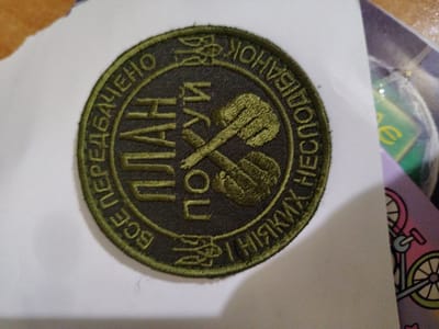A circular, olive-green embroidered patch is displayed on a white surface. The patch features Cyrillic text and a central emblem of two crossed fists superimposed on two crossed rifles. The text, in yellow thread, circles the emblem. The top of the patch reads "ПЕРЕДБАЧЕНО", and continues with "ПЛАН ПОГОДИ / З НІЯКИХ", and concludes with "ВСЕ". The emblem in the center contains two raised fists and what appears to be a stylized trident or a similar national symbol. The patch is slightly curved, suggesting it is a fabric appliqué. The white surface beneath the patch has some light gray speckled text, possibly from another item or a printer. In the background to the right, a portion of a colorful object with a drawing of a bicycle and abstract shapes is visible. The lighting appears to be artificial and casts a subtle shadow to the left of the patch. The overall image quality is slightly grainy. The location in Okhtyrka, Ukraine, is not visually indicated by any specific landmarks or contextual cues within the image itself.