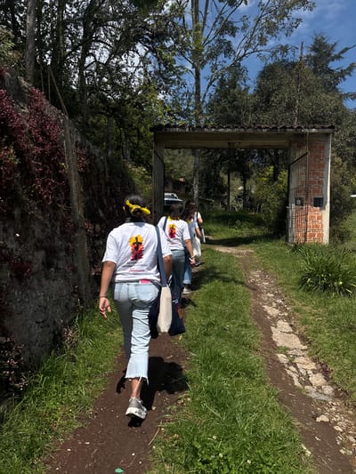 A medium shot shows a dirt path cutting through a grassy area in San Francisco Zacapexpan, Mexico. Three women wearing white t-shirts with a colorful graphic design on the front walk along the path, away from the viewer. The women are fair-skinned and wearing light blue jeans. The first woman is wearing a yellow bow in her hair, and a beige bag is slung across her shoulder. A stone wall and trees line the left side of the path. The right side is mostly green grass. A brick and concrete structure spans the path ahead of the women.