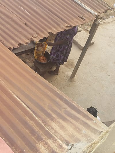 In Alalubosa, Nigeria, an overhead shot depicts a person wearing a patterned purple garment pouring contents from a yellow bag into a black bucket. The scene is situated on corrugated metal roofing panels interspersed with segments of concrete and a wooden beam. Another individual with dreadlocks is visible near the edge of the roof.