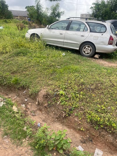 The image captures an outdoor scene dominated by a light-colored sedan car, appearing abandoned or in disrepair, set against an overgrown landscape in what is likely a semi-urban or rural area, consistent with Osogbo, Nigeria.
The primary subject is a silver or white sedan, heavily damaged and aged. It features a roof rack and shows signs of rust and faded paintwork. The rear driver's side tire (as oriented in the rotated image) is visible and appears flat or deflated, while the front passenger-side wheel is completely missing, exposing the wheel well. Another darker vehicle is partially visible behind the sedan.
The car is situated on a patch of uneven ground covered with a mix of tall green grass and sparse vegetation, interspersed with reddish-brown dirt. Small pieces of litter, including plastic bags, are scattered throughout the ground. In the background, dense green trees and bushes frame the scene. One of the cropped views reveals an unfinished or under-construction building with exposed concrete in the distance, alongside other structures, indicating a developing locale.
The scene appears to be set during daytime under an overcast sky, as no direct sunlight or clear shadows are visible. There are no people present, and no specific activity is taking place, suggesting the car has been left in its current state. No legible text is discernible on the vehicle or in the environment. The overall impression is one of neglect or disuse in an unkempt outdoor setting.