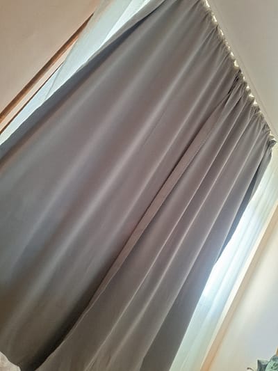 This image depicts an interior scene in Katina, Bulgaria, primarily featuring a window area covered by two layers of curtains. The foreground presents a grey, opaque fabric curtain, characterized by prominent vertical folds and pleats along its length. This curtain is suspended from a rod, indicated by gathered fabric and attachment points visible at the top. Behind the grey curtain, a sheer, white or off-white fabric curtain is visible, allowing diffused natural light to filter into the space. A strong light source, consistent with a window, is present behind both curtain layers, casting bright illumination from the upper left and right edges of the frame where the curtains are not fully closed. A segment of a light-colored window frame or interior wall trim is partially visible along the upper left margin of the image. The ceiling or upper wall area, appearing light-colored, is visible above the curtain rod. No individuals, actions, or specific objects beyond the curtains and surrounding architecture are discernible.