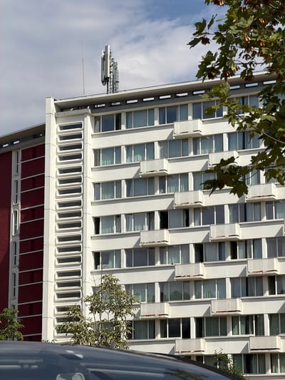 The image captures a side view of a modern building with a distinctive facade. The building is tall and rectangular, with a combination of white and dark red sections. The white section of the building is characterized by repeating horizontal bands of windows and balconies, creating a stepped effect. Each balcony is shallow and enclosed by white railings. A prominent feature of the white section is a vertical column on the left side, adorned with a series of narrow, rectangular, recessed windows.

The dark red section of the building, on the left, presents a plainer appearance with horizontal rows of windows that are set further back and appear smaller than those on the white section. It also has recessed vertical elements that resemble windows but are less distinct.

At the top of the white section, a telecommunications antenna is visible. The building is partially obscured by a large tree in the foreground, on the right side, with green leaves and some dried seed pods hanging from its branches. A portion of a dark-colored car is visible at the very bottom of the image, suggesting it is parked in front of the building. The sky above is partly cloudy, with patches of blue sky interspersed with white clouds. The lighting indicates it is daytime.

This photograph was taken in Antony, France, specifically at 12 Rue des Glaïeuls.