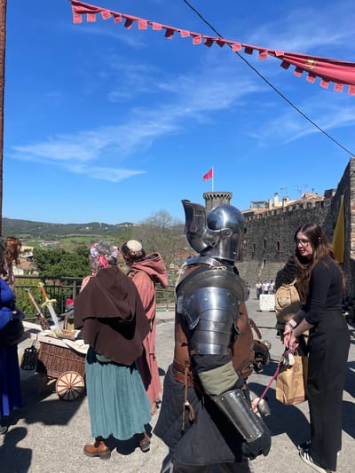 The image depicts a scene in Hostalric, Spain, characterized by a medieval reenactment or fair. A person dressed in full medieval armor stands prominently in the foreground, with a woman in black clothing adjusting something near the armor. Behind the armored figure are other individuals in medieval attire, including one in a brown robe, suggesting a monk or friar. A woman wearing a flower crown is visible, standing close to swords and a basket. In the background, the stone walls and towers of the Castle of Hostalric are visible. Red banners hang along the side, and the clear blue sky with scattered clouds serves as the backdrop. The landscape reveals buildings and green fields beyond the castle, which marks the general vicinity of Hostalric.