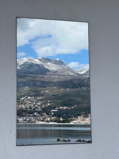 The image shows a reflective surface, potentially a mirror or a glossy print, reflecting a landscape. The reflected scene depicts a mountain range with visible snow cover on the upper slopes. Below the mountains is a densely populated town or city situated along a body of water. The sky is blue with scattered clouds. The town appears to have a mix of residential and commercial buildings. Rocks appear to be placed on the shoreline.
There is no explicit mention of "Provodina" in the image; however, the landscape is potentially reflecting the Bay of Kotor, Montenegro.
The image depicts landscape photography and nature reflecting in the glass.