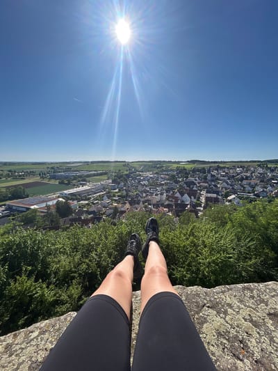 A high-angle, daytime, outdoor shot features a person's legs and feet dangling over the edge of a rocky precipice. The person is wearing black athletic shorts and black sneakers. The lower half of their legs is visible, with their feet dangling in the air. Below them is a lush green area filled with trees and vegetation. Beyond this green area is a view of the city of Untergruppenbach, Germany, characterized by densely packed buildings with varying roof colors, interspersed with trees and greenery. In the distance, the city transitions into agricultural fields. The sky above is a clear, bright blue, with a prominent sunburst in the upper left corner, casting a lens flare across the image. A faint contrail is visible in the sky near the sun. The rock surface appears to be light-colored with lichen or moss growth.