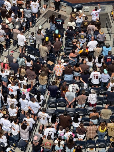 This image shows a large group of people sitting in stadium seats at a baseball game. Most people are wearing baseball jerseys, with many of the jerseys having "GWYNN" and the number 19 on the back. The name "TATIS JR" is also visible on a number of jerseys. Other names include "GALLEGO", "FRANCE", "DHAIGHT", "MOLINI", "MACHADO", "HA-SEONG", and "HOUSSE". The baseball stadium appears to be in San Diego, United States, based on the Padres jerseys.