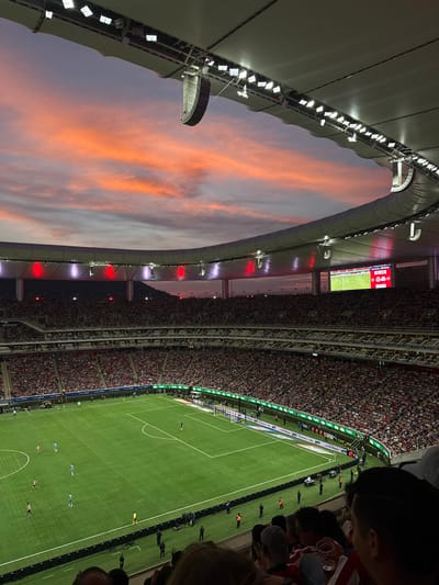 The image depicts a soccer game at Estadio Akron, a stadium located in Zapopan, Mexico. The stadium is packed with spectators. The soccer field is green, and multiple soccer players are on the field during the game. A large stadium screen displays the game information and time. The sky is visible through the stadium's open roof, showing a colorful sunset. The stadium structure includes lights and architectural features. People are visible in the foreground, suggesting the perspective is from within the stadium seating.