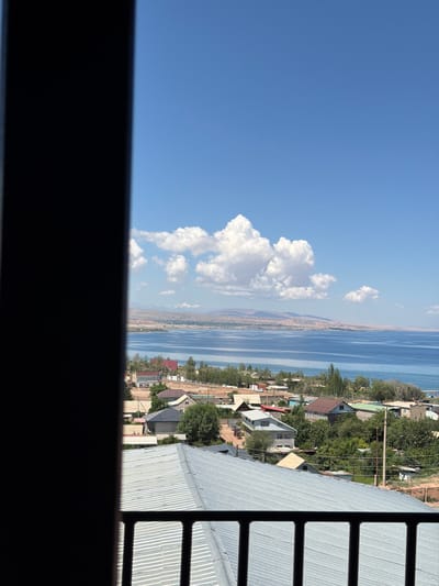 This is a high-angle, wide shot of a town with a body of water and mountains in the background. The foreground is dominated by a dark, out-of-focus vertical element on the left, likely a window frame or railing. In front of the body of water, a town is visible with a variety of houses, some with tin roofs and others with tile roofs. Trees are scattered throughout the town. The body of water appears to be a large lake or sea, with a distant shoreline and mountains beyond. The sky is bright blue with scattered white cumulus clouds. The scene is likely Kaji-Say, Kyrgyzstan, given the geographical features and architectural style. There are no people or animals visible in the image.