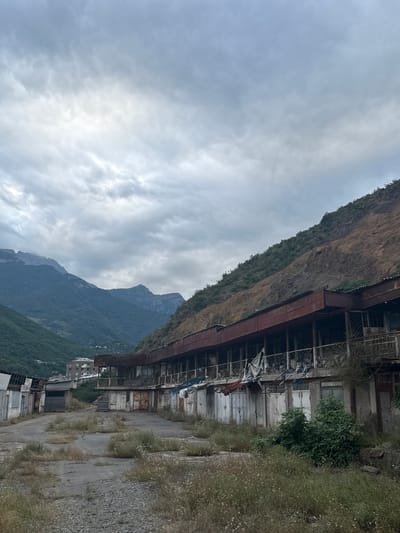 The image depicts an exterior view of a dilapidated building structure nestled against a steep, vegetated mountainside. The building appears to be multiple stories high with exposed framework and weathered facades. Some windows are visible, some boarded up, while others are open. Deteriorated balconies or walkways are present, adorned with what appear to be draped fabrics or tarps. Vegetation, including dense green foliage and dry grasses, grows around and on parts of the structure, suggesting abandonment or neglect. A gravel path runs alongside the building.

In the background, imposing, lush green mountains rise dramatically against a cloudy sky. The clouds are heavy and grey, indicating overcast or potentially rainy weather. The overall atmosphere is one of decay and natural reclamation. There are no people visible in the frame, and no text is discernible. The scene suggests a remote or possibly abandoned industrial or residential area, with the natural landscape dominating the man-made structure. The angle of the shot, looking upwards towards the building and mountains, emphasizes their scale and the ruggedness of the location.