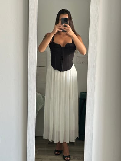 A woman is taking a mirror selfie in what appears to be a bedroom in Torremolinos, Spain. She is wearing a black corset top with a sweetheart neckline and a long, white pleated skirt. Her hair is long and dark, and she is holding a smartphone in her right hand to capture the reflection. Her left arm is also visible, and a tattoo can be seen on her upper arm. She is wearing black open-toed heels. The mirror is tall and framed with a textured white border. In the background, a wall with subtle texture is visible, along with a dresser with multiple drawers. To the right of the mirror, a dark green yoga mat is standing upright. The floor is a light-colored wood laminate.