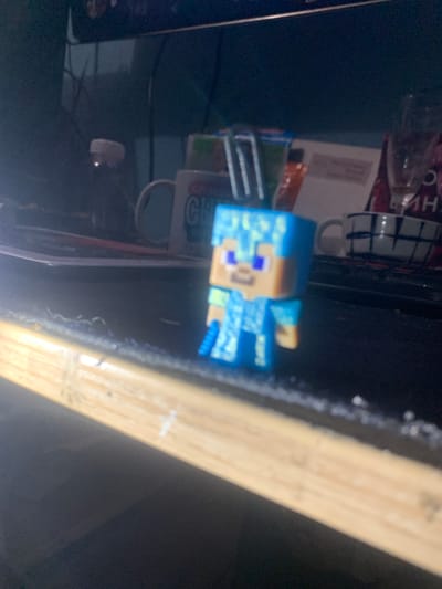A blurry, close-up shot captures a small, light blue Minecraft action figure in focus. The figure is made of plastic and appears to be a character from the game, with blocky features and pixelated designs. It has a light brown face with blue eyes and a light blue body adorned with intricate patterns in various shades of blue and white. The figure is positioned at an angle, with its left side closer to the camera.
Behind the action figure, the scene is dark and somewhat cluttered. To the left of the figure, there are hints of other objects, possibly on a shelf or desk, including what looks like a white box with some writing and a red and white checkered mug or container. There are also some blurry green and orange items. The background consists of a dark surface, likely a table or desk, with a wooden plank running diagonally across the right side of the frame. The plank is light brown and appears to be part of the furniture or structure in the setting. The overall lighting is dim, suggesting an indoor environment, potentially at night or in a poorly lit room. No specific environmental cues like weather or time of day are discernible beyond the indoor setting. The image is very shallow in depth of field, with most of the background and foreground out of focus.