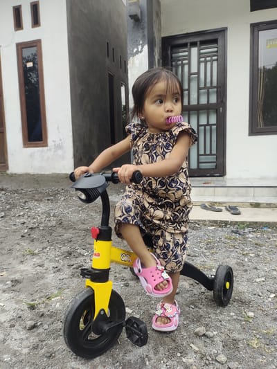 A young girl is standing on a yellow tricycle in front of a residential building. The girl is wearing a patterned beige and navy blue outfit and pink sandals adorned with cartoon characters. She has a pink object in her mouth that appears to be a pacifier or chew toy. The tricycle is yellow with black wheels and handlebars. The background features a building with white and dark grey walls, and a dark brown door. The ground is covered in gravel and dirt, with some patches of dry grass. The scene appears to be outdoors, likely in a yard or driveway, consistent with the residential setting of Cintadamai, Indonesia.