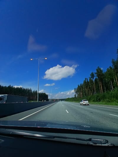 The image depicts a view from inside a vehicle traveling on a multi-lane highway. The sky is a vibrant blue with scattered white clouds. On the left side of the highway, a concrete barrier separates the lanes from a wooded area. A large white truck is visible behind the barrier. Tall streetlights with illuminated bulbs line the highway. On the right side of the road, a dense forest of pine trees stretches along the horizon. A white car is driving in the right lane. The road surface is asphalt with white lane markings. The foreground shows the interior of the vehicle, including the dashboard and windshield wipers. The scene appears to be a typical highway environment, possibly in a rural or suburban area. The location is specified as the city of Khrustal'nyy, Russia, suggesting this highway is situated in or near that city. There are no people visible in the image, other than occupants of the vehicles. The weather appears to be clear and sunny.