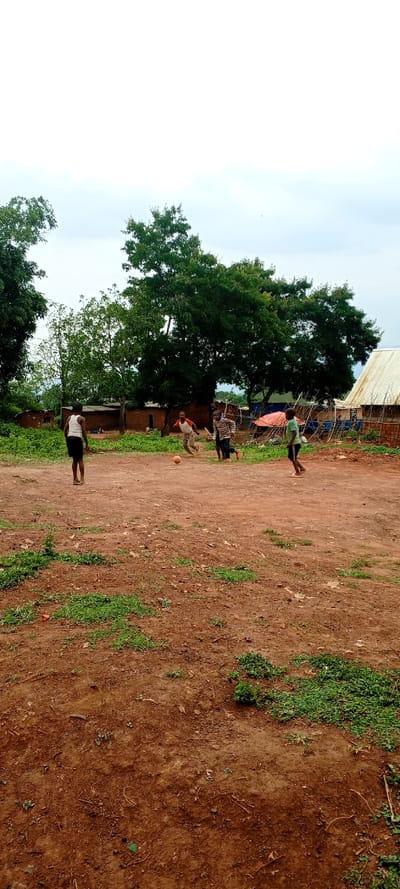 The image depicts an unpaved outdoor area in Kundu 1, Democratic Republic of the Congo, under an overcast sky. Five individuals, appearing to be children, are present.

In the mid-ground, four children are actively engaged in playing football with an orange ball. One child, wearing a white tank top and dark shorts, stands on the far left, facing away from the camera. To the right of the center, a child in an orange and white shirt and light pants is positioned near the orange football. Behind this child, another child in a striped shirt and dark pants runs towards the left. A fourth child, wearing a green shirt and dark shorts, runs on the far right of the playing area. A fifth individual is partially obscured in the background, near a structure on the left.

The ground surface consists primarily of reddish-brown earth, uneven in topography, with scattered patches of green vegetation and small debris. The background features several structures: unpainted brick or mud-brick buildings are visible on the left side, some appearing partially damaged. On the right, a building with a corrugated metal roof is present. A red tarpaulin or similar covering is visible in the center-background. Numerous large trees with dense green foliage are distributed across the mid-ground and background. The sky is uniformly light grey, indicating an overcast condition.
