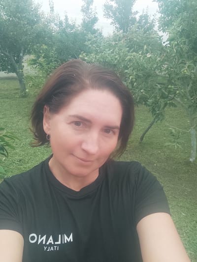 A mid-shot, selfie-style photograph captures a woman with shoulder-length brown hair and light skin. She is wearing a black t-shirt with white lettering that reads "MILANO YJATI". A small silver hoop earring is visible in her left ear. She is smiling slightly and looking directly at the camera. The background consists of a grassy area with several trees and green foliage, suggesting an outdoor setting, likely in Kobeliachok, Ukraine. The lighting is overcast.