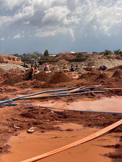 The image depicts a group of people working in a muddy, reddish-brown terrain, likely a construction or excavation site. The scene is oriented vertically, suggesting the original photo was taken sideways. The subjects are individuals engaged in manual labor, some appearing to be digging or moving earth. Several are dressed in casual clothing, with one person wearing a pink garment.
Objects in the scene include numerous pipes of varying colors, predominantly blue and orange, lying on the ground. Some pipes are submerged in the muddy water. The ground itself is a mixture of soil, rocks, and water, indicating a wet and possibly recently rained-upon environment. In the background, a rocky hillside rises, with some vegetation and buildings visible at the top, suggesting an inhabited area or a nearby town. The sky is partly cloudy, with patches of blue visible.
The activity appears to be related to earthmoving or construction work, possibly involving water management due to the presence of standing water and pipes. The time of day seems to be daytime, given the illumination and the sky conditions. The location cues, such as the reddish soil and the style of buildings in the background, align with the provided context of Reifild-Ist, Nigeria. The overall impression is one of arduous manual labor in a challenging environment. No visible text is present in the image.