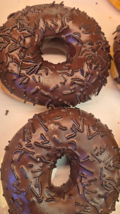 Two chocolate-frosted donuts with chocolate sprinkles are visible in the foreground of the image. The donuts are round with a hole in the center and are placed on a light-colored surface. The top donut is closer to the viewer and is in focus, while the bottom donut is partially out of focus. A third donut is partially visible in the background on the right side of the image. The lighting is bright and even, highlighting the glossy texture of the frosting and the individual sprinkles. There is no information available to indicate the location of this image within the city of Preiļi, Latvia.