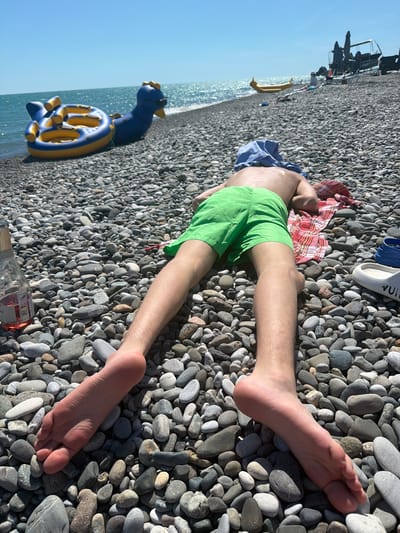 In Amzara, Abkhazia, a person is lying face down on a pebble beach, with their head covered by a blue cloth. They are wearing green shorts and are resting on a red and white checkered towel. To their left, a bottle of rosé wine stands upright. In the background, inflatable water toys, including a blue dragon-shaped one and a yellow ring float, are visible near the water's edge. Further along the beach, there are lounge chairs, umbrellas, and other beachgoers. The sun is shining brightly, and the sea is calm.