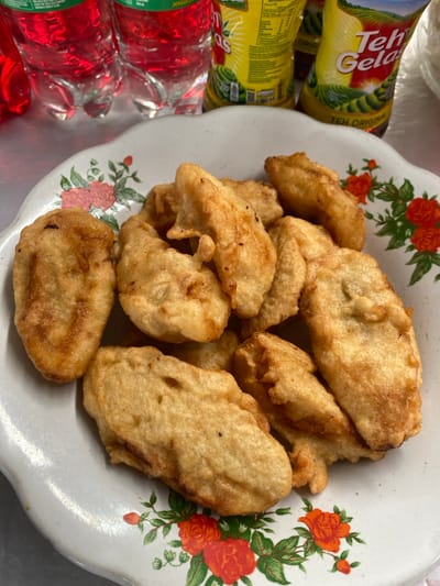 A white, scalloped-edge plate with a floral pattern featuring red and pink roses and green leaves occupies the foreground and center, holding approximately fifteen to twenty irregular-shaped, golden-brown deep-fried battered food items, consistent with Indonesian pisang goreng (fried bananas). The plate rests on a light-colored, possibly metallic, surface. In the background, from left to right, two clear plastic bottles containing red liquid are visible. Adjacent to these are two yellow stand-up pouches clearly labeled "Teh Gelas" and "TEH ORIGINAL." No individuals are present, and no specific actions or interactions are depicted. The visible branding for "Teh Gelas" identifies the beverages as common Indonesian products. While the scene indicates an Indonesian context, the precise location within Jepara is not discernible from the visual evidence.