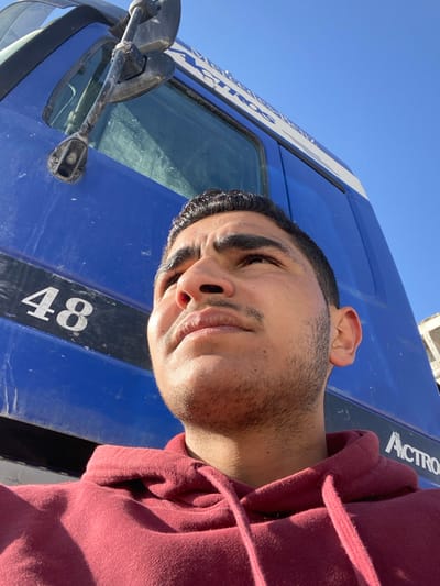 A man in a maroon hoodie poses beside a large blue truck on a sunny day. The truck has "Actros" written on its side. Part of the truck's window and side mirror are visible. The man's face is close to the camera, showing his features like his hairline and stubble. The angle of the photograph is slightly tilted, giving a sense of a selfie.