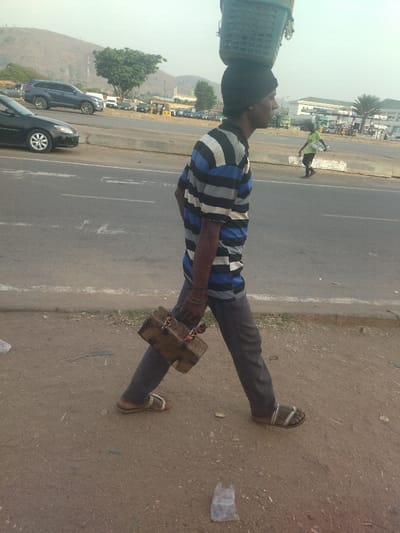 A man is walking on the shoulder of a paved road in the city of Maraba, Nigeria, carrying a large wooden box in his right hand and a green plastic basket on his head. He is wearing a black hat, a blue and white striped shirt, gray pants, and sandals. Another man wearing a green t-shirt is walking further down the road. The road is empty of traffic, and the environment is dry and dusty.