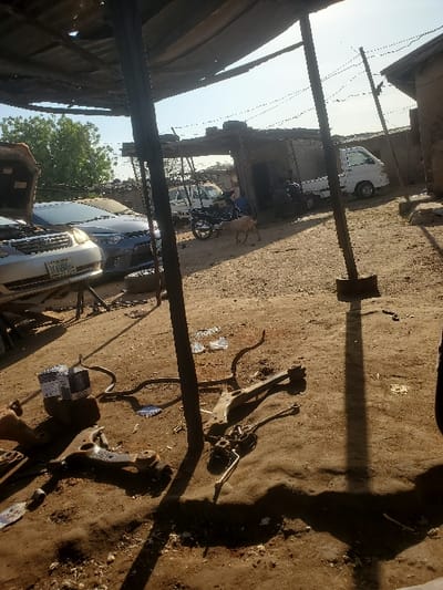 The image displays an outdoor automotive repair area in Kaduna South, Nigeria, under clear, bright daylight. In the left foreground, a silver sedan with an open hood rests on a jack stand, positioned ahead of a dark grey sedan also featuring an open hood. The unpaved dirt ground in the foreground and midground is littered with various discarded automotive components, including suspension control arms and linkages, alongside two white boxed items. Overhead, a rudimentary canopy structure, supported by dark metal poles, provides partial shade. In the midground, a black motorcycle is parked near a white mini-pickup truck, behind which another white vehicle is partially visible. Two individuals are seated or crouching in the midground, near the motorcycle and truck. A light-colored goat walks rightward across the midground. The background consists of several single-story structures appearing to be constructed from concrete or mud bricks.