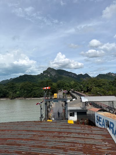 A ferry terminal is depicted with a body of water to the left, lush green trees and rocky mountains in the background, and a cloudy sky above. In the foreground, a rusty corrugated metal roof is visible. The ferry terminal itself has a ramp leading to a covered walkway. A windsock, a yellow rectangular object, and flags are present on the terminal. Several people are visible on the ramp and walkway, some appearing to be passengers and others possibly staff. A white shipping container with blue lettering, reading "SEAR," is also positioned at the terminal. The scene appears to be a functional ferry terminal in a tropical or subtropical region, given the vegetation and mountainous landscape.