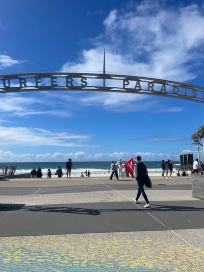 The image depicts the Surfers Paradise sign in Gold Coast, Australia. The sign is an archway with metal lettering spelling out "SURFERS PARADISE". Beneath the sign, people are visible on a beach and a paved walkway. The ocean is in the background with waves and a few sailboats. The sky is blue with scattered clouds. Several individuals are walking and standing, with some holding flags. The pavement has a patterned design.
