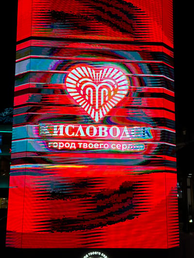 The image shows a large, vertical digital advertisement displayed on the side of a building in Kislovodsk, Russia. The advertisement is predominantly red with a stylized heart graphic at the top, containing white stylized letters. Below the heart, text in white Cyrillic letters reads "КИСЛОВОДСК" and "город твоего сердца" (Kislovodsk, the city of your heart). The digital display exhibits significant visual distortion, with horizontal banding and glitching effects, suggesting a technical issue or an intentional artistic choice. The background is dark, indicating it is likely nighttime. The building appears to be a modern structure with segmented panels that are illuminated by the advertisement.