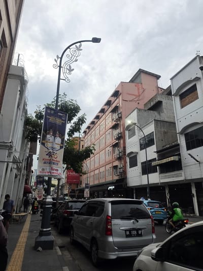 The image depicts a street scene in the city of Medan, Indonesia. A light gray sedan is parked on the right side of the street, facing away from the camera. Behind it, a blue car is driving away, and a motorcyclist in a green jacket is also moving away. The street is lined with buildings of various architectural styles and colors, including a pink, multi-story building and white buildings with tiled facades. A lamppost with decorative metalwork stands prominently in the foreground, displaying a banner. The banner appears to be a celebration of "HUT 435 KOTA MEDAN" which translates to "435th Anniversary of the City of Medan." There are also some pedestrians visible on the sidewalk on the left side of the street, and more parked cars. The sky is overcast with clouds. The scene suggests a typical urban environment with commercial and residential buildings, traffic, and public decorations.