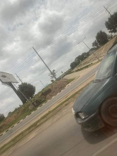 The image shows a street scene in Kuchingoro, Nigeria, with a car partially visible in the foreground. The road is adjacent to a sidewalk with a low wall, followed by green vegetation. Streetlights are visible along the road. Utility poles with electrical lines run parallel to the road on the opposite side. A basketball hoop is also visible. The sky is overcast with heavy clouds.