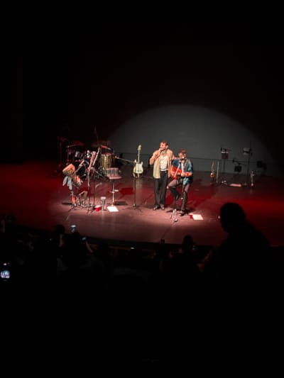 The image depicts a stage scene, likely from a performance or concert, in Tlalnepantla, Mexico. Two individuals are standing center stage under a spotlight. A band's musical instruments, including a drum set and a guitar on a stand, are visible behind them. A blurred audience is visible in the foreground, with some individuals holding up phones. The lighting is dim, with a focused spotlight on the performers and the stage floor illuminated with red light.