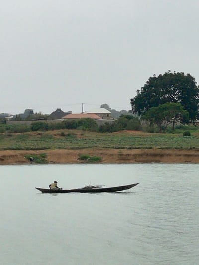 A man in a narrow boat is on the water, next to a grassy bank and some buildings, in Nabatum, Nigeria.